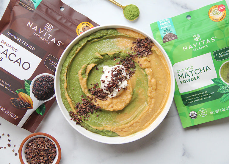 A bowl of swirled green and orange smoothie bowl topped with coconut cream and cacao nibs, surrounded by packages of Navitas Organics Cacao Nibs and Matcha Powder.