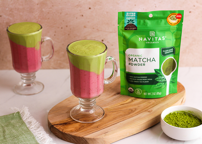 Two layered smoothies in clear mugs beside a package of Navitas Organics Matcha Powder, displayed on a wooden board against a neutral background.