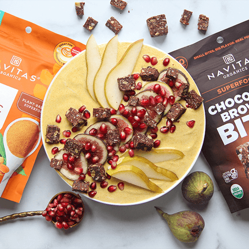 A yogurt bowl garnished with pears, pomegranate seeds, and Chocolate Brownie BITES, accompanied by a spoonful of pomegranate seeds and packages of Navitas Organics Turmeric Powder and Chocolate Brownie BITES in the background.