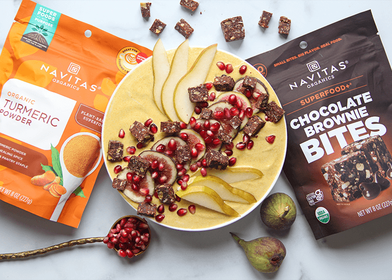 A yogurt bowl garnished with pears, pomegranate seeds, and Chocolate Brownie BITES, accompanied by a spoonful of pomegranate seeds and packages of Navitas Organics Turmeric Powder and Chocolate Brownie BITES in the background.