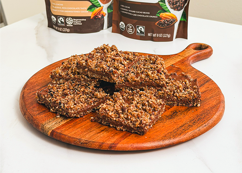 Homemade chocolate pecan bars on a wooden serving board in front of bags of Navitas Organics Cacao Powder and Cacao Nibs.