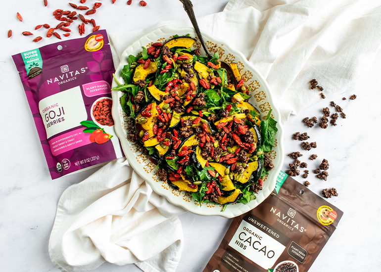 Colorful salad with arugula, acorn squash slices, goji berries and candied cacao nibs on a plate, alongside packages of Navitas Goji Berries and Cacao Nibs.
