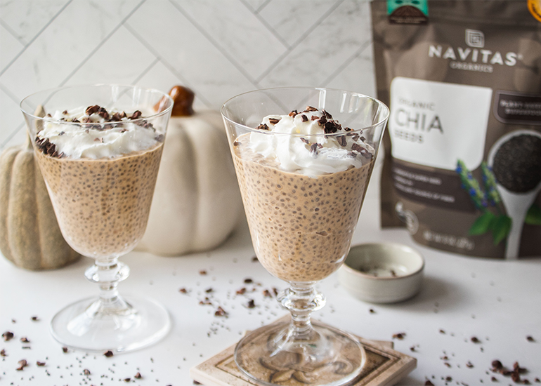 Two glasses of chia pudding topped with whipped cream and cacao nibs, displayed on a kitchen counter alongside a bag of Navitas Organics Chia Seeds.