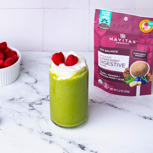 A vibrant green smoothie topped with whipped cream and raspberries, served next to a bowl of raspberries and bananas, with a package of Navitas Organics Superfood+ Digestive Blend visible on the counter.
