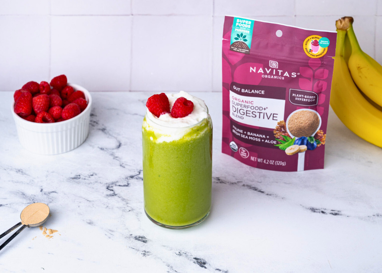 A vibrant green smoothie topped with whipped cream and raspberries, served next to a bowl of raspberries and bananas, with a package of Navitas Organics Superfood+ Digestive Blend visible on the counter.