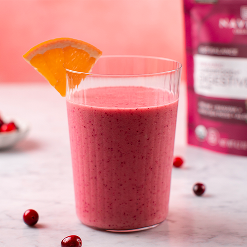 A smoothie in a clear glass garnished with an orange slice, with a bag of Navitas Organics Superfood+ Digestive Blend and some scattered cranberries in the background. 