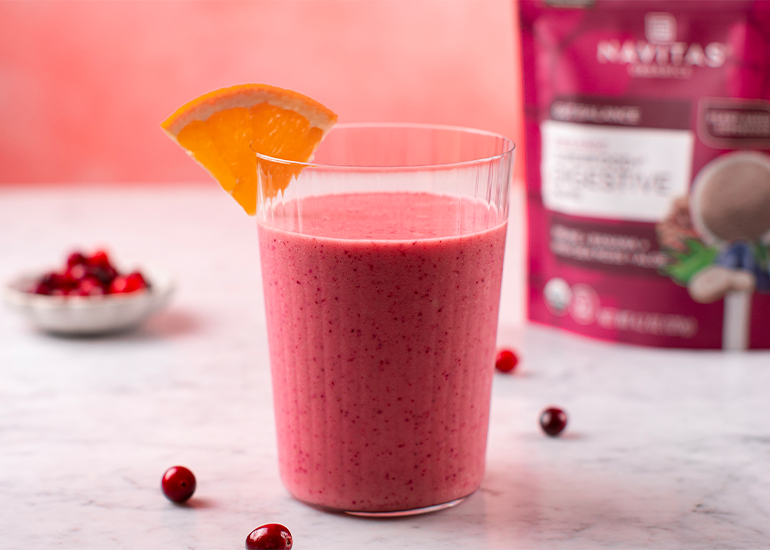 A smoothie in a clear glass garnished with an orange slice, with a bag of Navitas Organics Superfood+ Digestive Blend and some scattered cranberries in the background. 