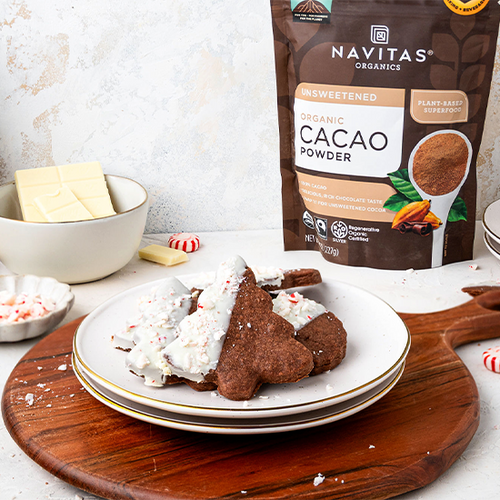 A festive dessert plate featuring chocolate cookies half-dipped in white chocolate, sprinkled with crushed candy canes, accompanied by a bowl of white chocolate and a package of Navitas Organics Unsweetened Cacao Powder on a decorated holiday table.