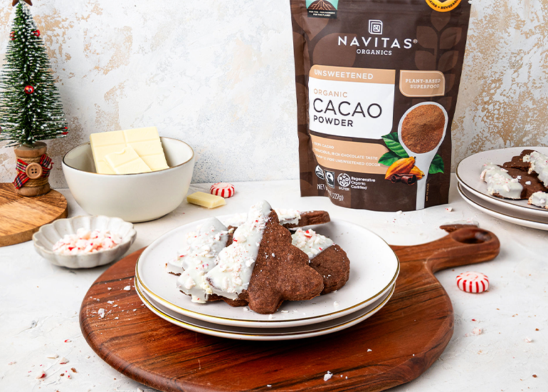 A festive dessert plate featuring chocolate cookies half-dipped in white chocolate, sprinkled with crushed candy canes, accompanied by a bowl of white chocolate and a package of Navitas Organics Unsweetened Cacao Powder on a decorated holiday table.