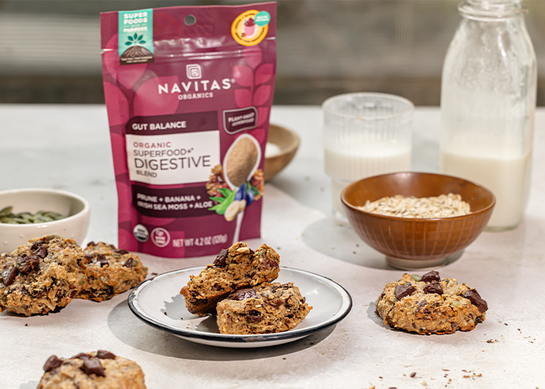 A display of freshly baked cookies on a plate beside a bag of Navitas Organics Superfood+ Digestive Blend. Ingredients like oats and a jug of milk are visible in the background on a kitchen counter.