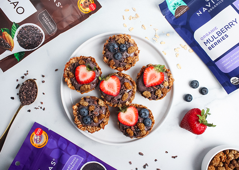 A plate of 6 oatmeal cups filled with yogurt mixed with Navitas Organics Acai Powder, topped with sliced strawberries, blueberries, and Navitas Organics Mulberries and Cacao Nibs, surrounded by bags of Navitas Organics products.