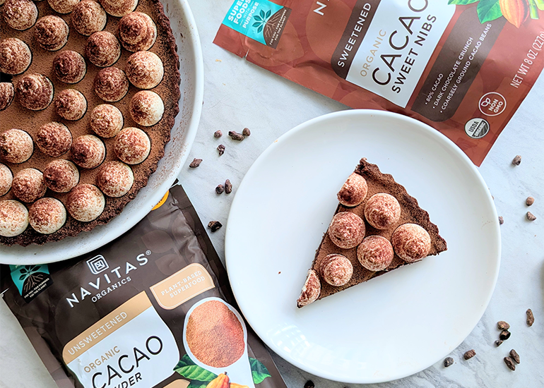 A chocolate cake decorated with dusted spheres on a white plate next to a slice on a separate plate, surrounded by bags of Navitas Organics Cacao Powder and Cacao Sweet Nibs.