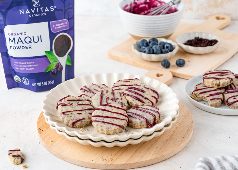 Shortbread cookies with a purple drizzle on a stack of white plates on top of a wooden cutting board, surrounded by bowls of blueberries and maqui powder, with a package of Navitas Organics Maqui Powder visible.