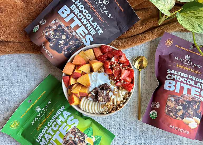 A variety of Navitas Organics products including Superfood+ Chocolate Brownie BITES, Salted Peanut Chocolate BITES and Lemon Cashew Matcha BITES, displayed around a bowl of yogurt topped with sliced fruits and granola.
