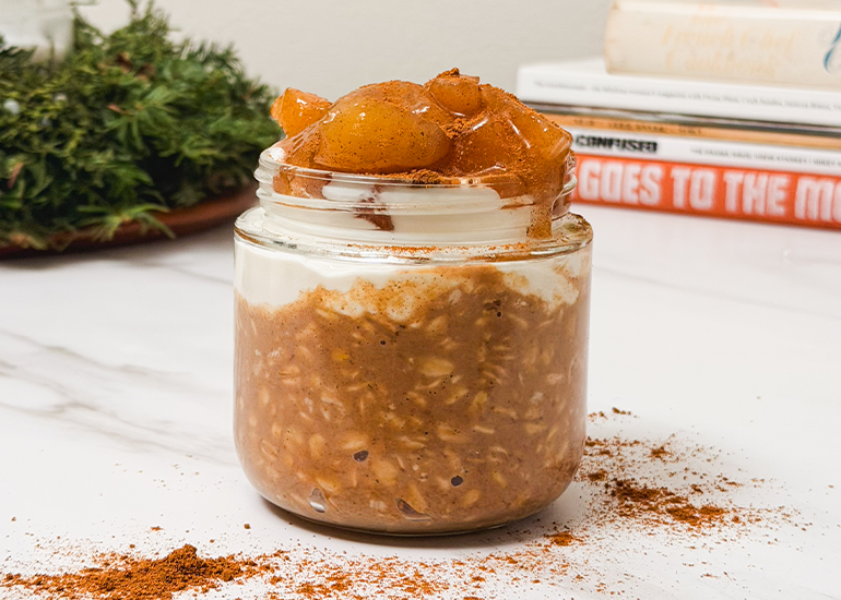 A jar of overnight oats made with Navitas Organics Superfood+ Digestive Blend topped with yogurt and apples on a marble countertop with books and plants in the background.