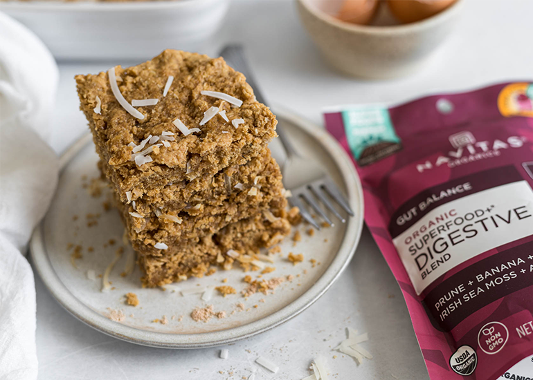 A stack of oat bars topped with coconut shavings on a ceramic plate, with a fork beside it. In the background, there are eggs and a bag of Navitas Organics Superfood+ Digestive Blend.