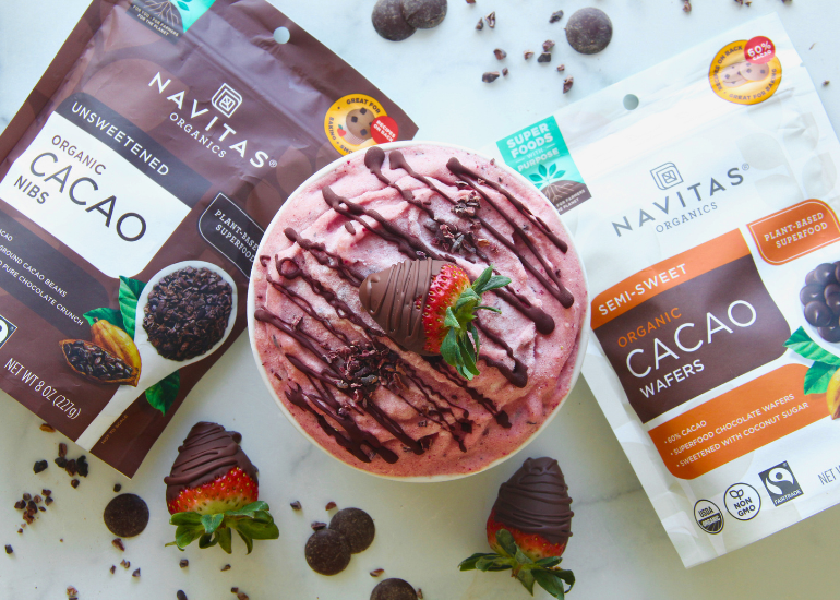 A strawberry smoothie bowl topped with a chocolate-covered strawberry and chocolate drizzle is flanked by packages of Navitas Organics Cacao Nibs and Semi-sweet Cacao Wafers. Additional chocolate pieces and strawberries are scattered around.