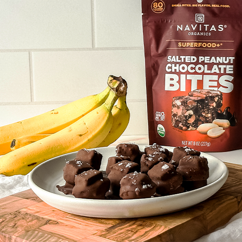 A plate with chocolate-covered snacks is in the foreground, sprinkled with sea salt. Nearby, a bunch of bananas rests on a wooden surface. Behind them, a bag of Navitas Organics Salted Peanut Chocolate Bites is displayed against a white tiled background.