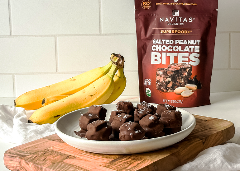 A plate with chocolate-covered snacks is in the foreground, sprinkled with sea salt. Nearby, a bunch of bananas rests on a wooden surface. Behind them, a bag of Navitas Organics Salted Peanut Chocolate Bites is displayed against a white tiled background.