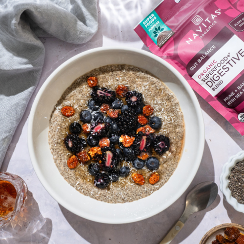A bowl of chia pudding topped with blueberries, blackberries, and goldenberries, accompanied by a bag of Navitas Organics Superfood+ Digestive Blend and a small bowl of chia seeds on a sunlit table.