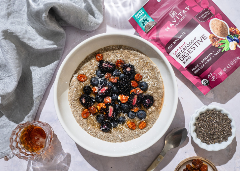 A bowl of chia pudding topped with blueberries, blackberries, and goldenberries, accompanied by a bag of Navitas Organics Superfood+ Digestive Blend and a small bowl of chia seeds on a sunlit table.