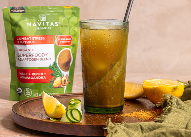 A tall glass filled with an adaptogenic mocktail surrounded by fresh lemon wedges and a bag of Navitas Organics Superfood+ Adaptogen Blend
