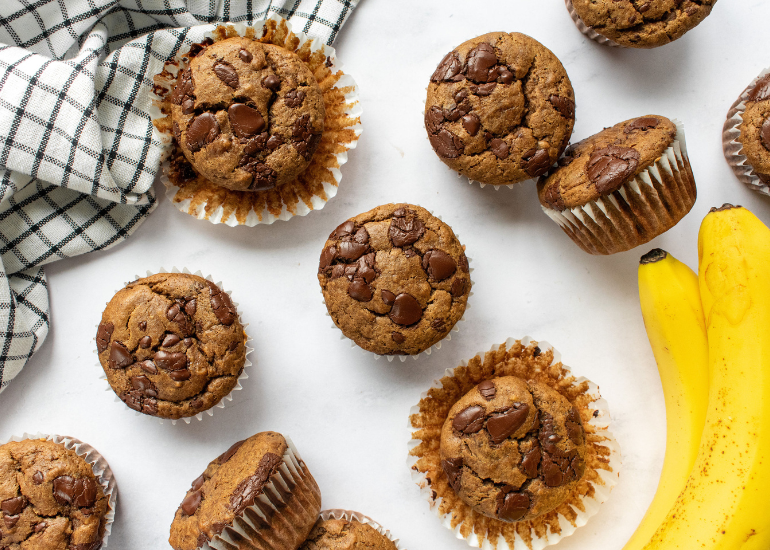 Chocolate studded banana bread muffins on a white surface surrounded by a checkered towel and fresh bananas.