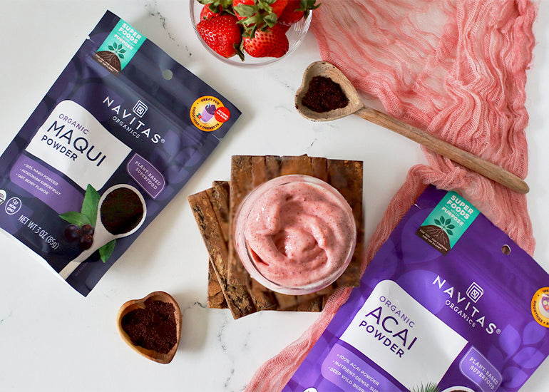 An overhead shot of a pink smoothie on a stack of wooden coasters surrounded by bags of Navitas Organics Maqui Powder, Acai Powder, wooden spoonfuls of the powders, a dish full of fresh strawberries and a pink linen cloth.
