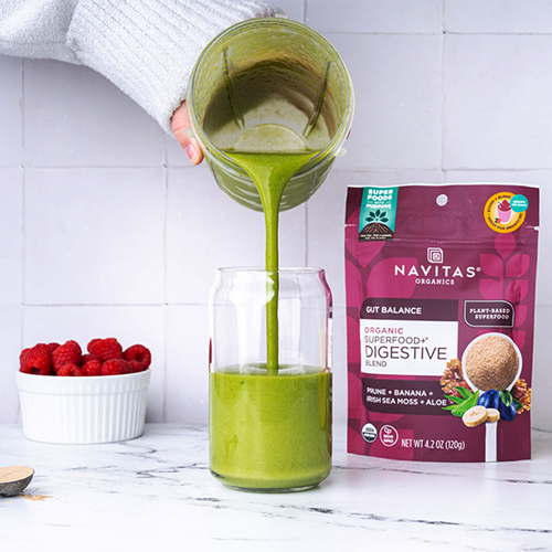 Person pouring a green smoothie into a glass next to a package of Navitas Organics Superfood+ Digestive Blend and fresh raspberries and bananas on a kitchen counter.