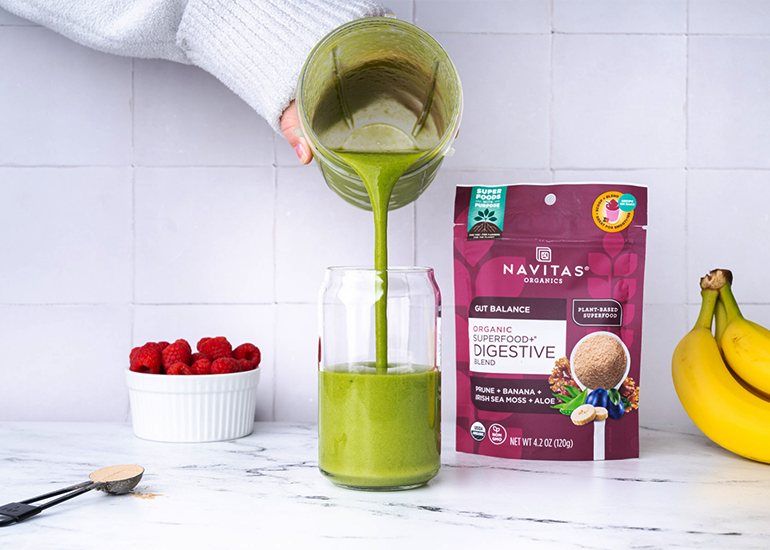 Person pouring a green smoothie into a glass next to a package of Navitas Organics Superfood+ Digestive Blend and fresh raspberries and bananas on a kitchen counter.