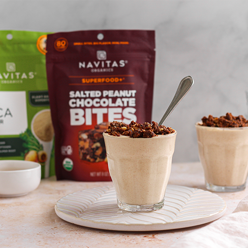 Two glasses filled with a creamy smoothie, topped with crumbled snacks and spoons inside. Behind them, packages of Navitas Organics Maca Powder and Salted Peanut Chocolate BITES are visible. A few empty bowls and glasses are on the side.