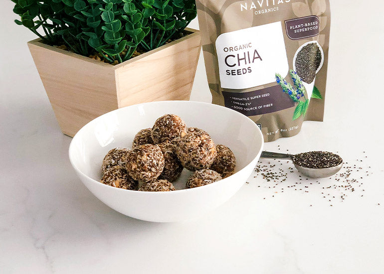 A bowl of chia bites made with Navitas Organics Chia Seeds in front of a plant, a bag of chia seeds and a scoop filled with chia seeds