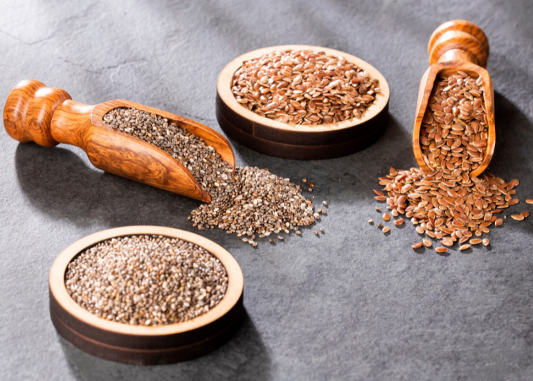 Two wooden spoons spilling out chia seeds and flaxseeds next to dishes filled with chia seeds and flax seeds on a grey marble counter.