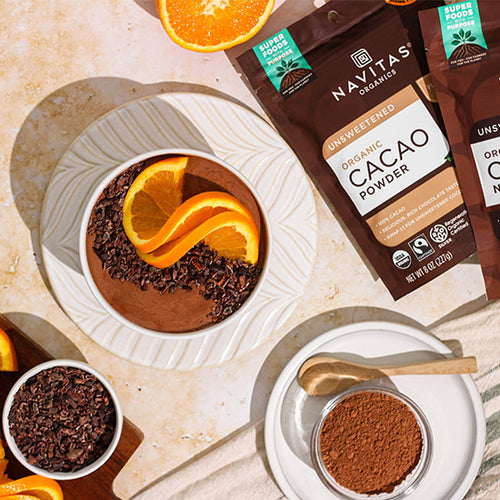 A chocolate orange smoothie bowl surrounded by Navitas Organics Cacao Powder and Cacao Nibs in dishes and in bags