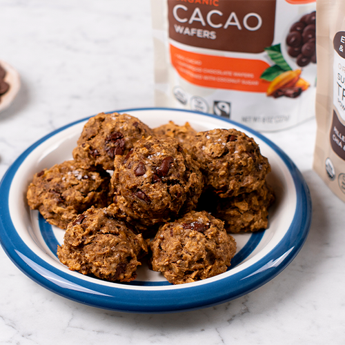A plate of cookie is placed on a marble countertop. Behind the plate are bags of Navitas Organics Semi-Sweet Cacao Wafers and Superfood+ Tri-Seed Powder Blend. A small dish of cacao wafers is visible on the side.