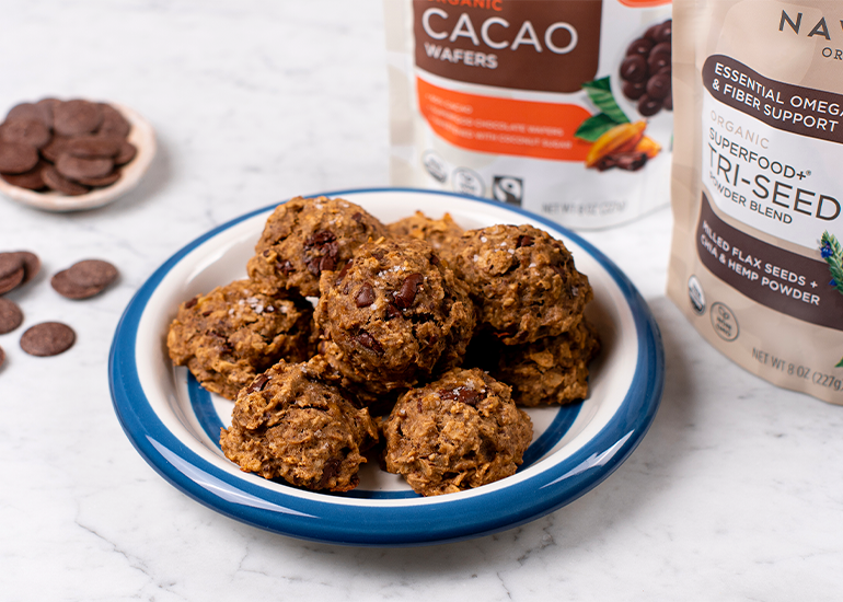 A plate of cookie is placed on a marble countertop. Behind the plate are bags of Navitas Organics Semi-Sweet Cacao Wafers and Superfood+ Tri-Seed Powder Blend. A small dish of cacao wafers is visible on the side.