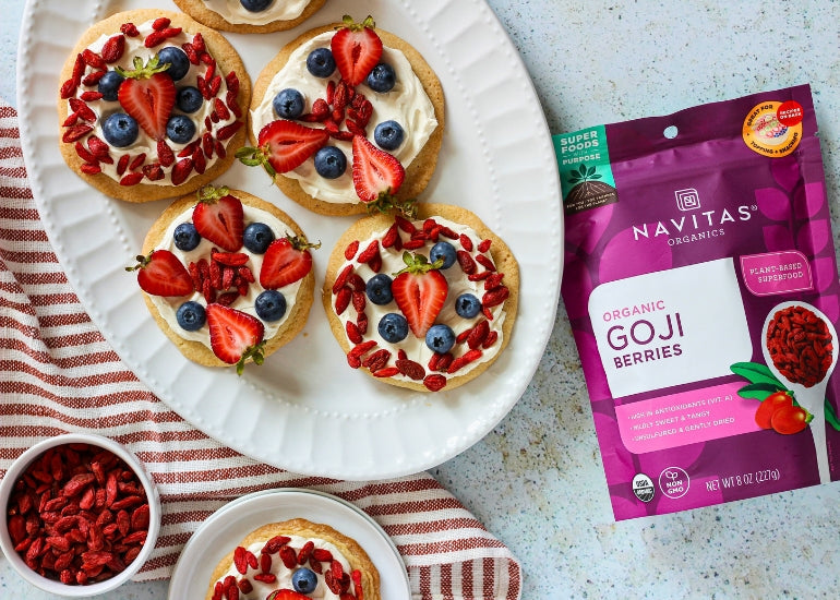 A plate of sugar cookies topped with cream cheese frosting, strawberries, blueberries and goji berries, displayed near a bowl of goji berries and a bag of Navitas Organics Goji Berries.
