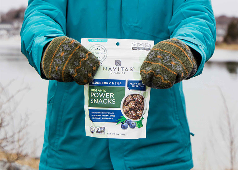 A woman in a winter coat and gloves holding a bag of Navitas Organics Blueberry Hemp Power Snacks.