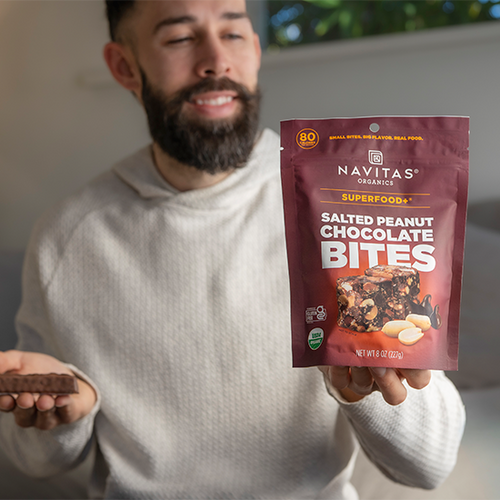 Person holding a package of Navitas Organics Superfood+ Salted Peanut Chocolate BITES in one hand and a chocolate-covered candy bar in the other, while looking at the BITES and smiling in a bright, cozy indoor setting.