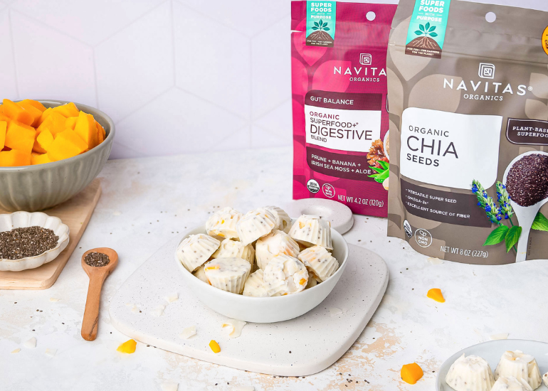 A kitchen scene featuring bags of Navitas Organics Superfood+ Digestive Blend and Organic Chia Seeds. A bowl of mango chunks, a small dish of chia seeds with a wooden spoon, and a plate of white snack cups are also visible on a light-colored counter.