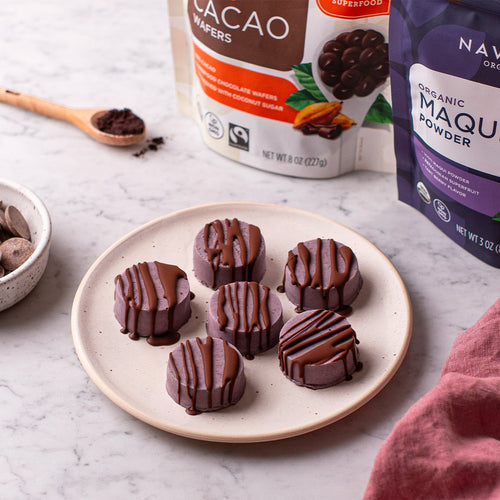 Plate of chocolate drizzled maqui treats next to a bowl of cacao wafers, with Navitas Organics Semi-sweet Cacao Wafers and Navitas Organics Acai Powder packages in the background.