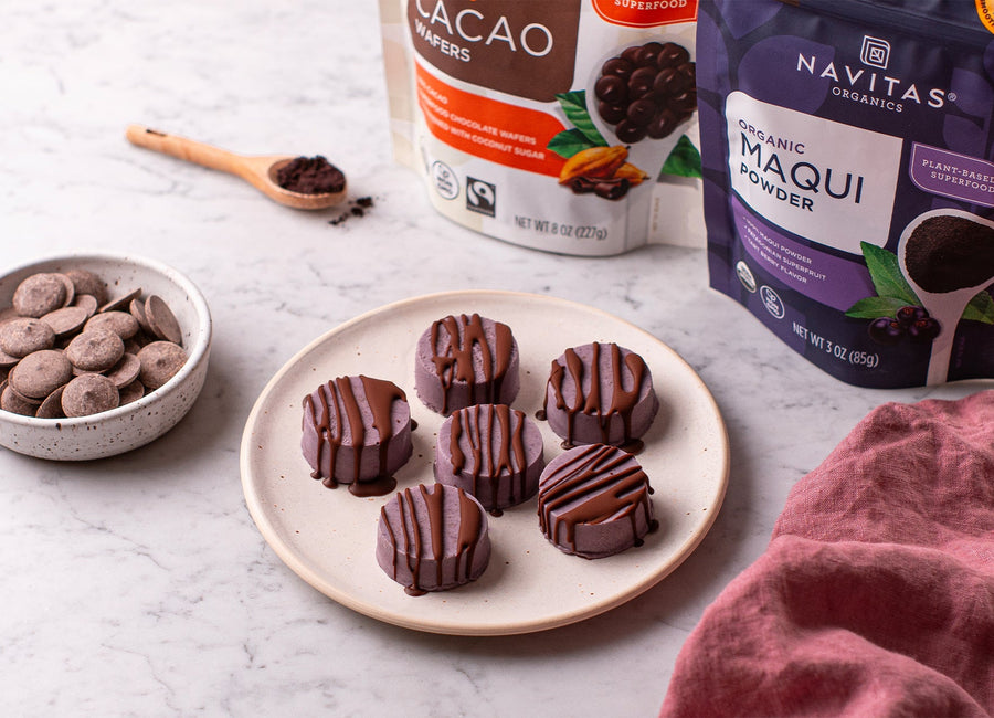 Plate of chocolate drizzled maqui treats next to a bowl of cacao wafers, with Navitas Organics Semi-sweet Cacao Wafers and Navitas Organics Acai Powder packages in the background.