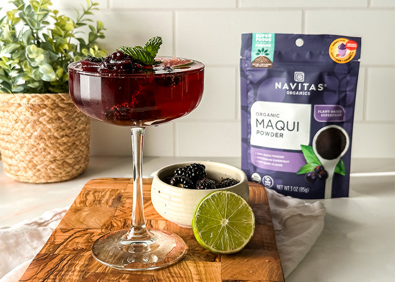A tall cocktail glass filled with a dark berry-colored mocktail, garnished with blackberries and fresh mint leaves, surrounded by a halved lime, a dish filled with blackberries and a bag of Navitas Organics Maqui Powder.