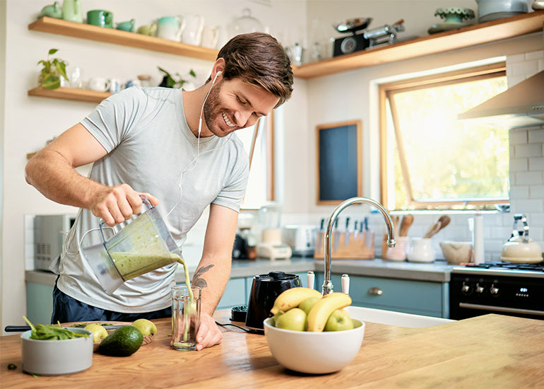 A man standing in a kitchen pouring a green smoothie from a blender into a tall serving glass