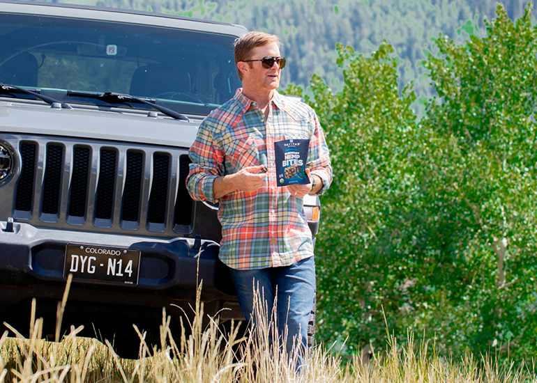 Person in sunglasses and plaid shirt standing next to a Jeep vehicle, holding a bag of Navitas Organics Superfood+ Blueberry Hemp BITES in a natural setting.