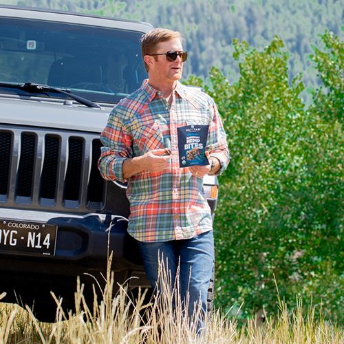 Person in sunglasses and plaid shirt standing next to a Jeep vehicle, holding a bag of Navitas Organics Superfood+ Blueberry Hemp BITES in a natural setting.