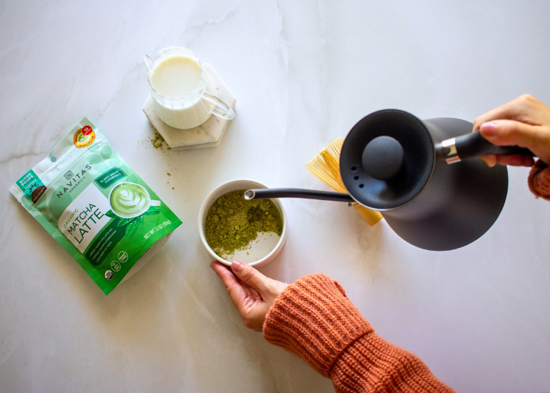 Person pouring hot water from a black kettle into a cup filled with matcha powder next to a package of Navitas Organics Matcha Latte Mix on a marble surface.