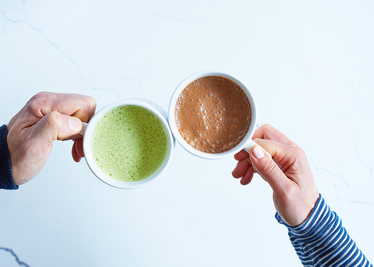 A man holding a matcha latte and a woman holding a hot chocolate, toasting each other