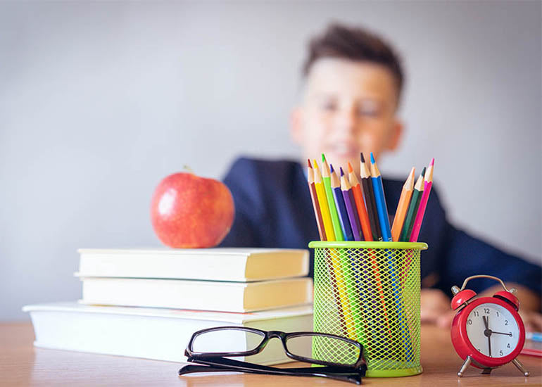 A student sitting at a desk with a stack of books, a container of colored pencils, an apple, a clock and reading glasses.
