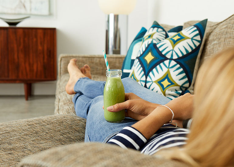 A woman laying on a couch holding a green smoothie made with Navitas Organics Maca Powder.